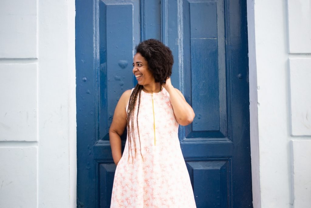 Ana Lira está de pé em frente a uma porta, sorrindo, com a mão no cabelo, de vestido, com a mão no bolso.